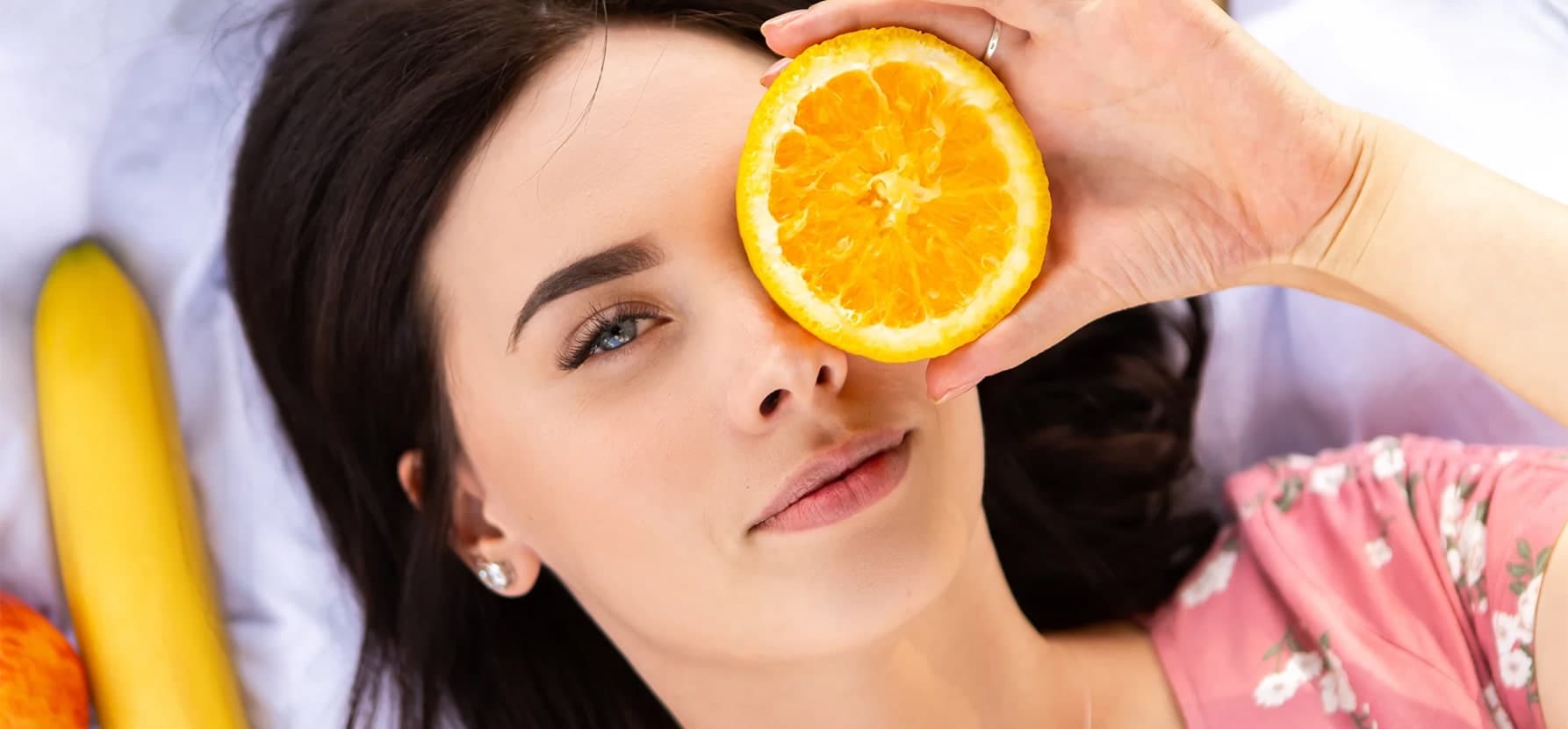 A woman holds an orange slice to her face, highlighting the benefits of Vitamin C for brightening skin and reducing dark spots.